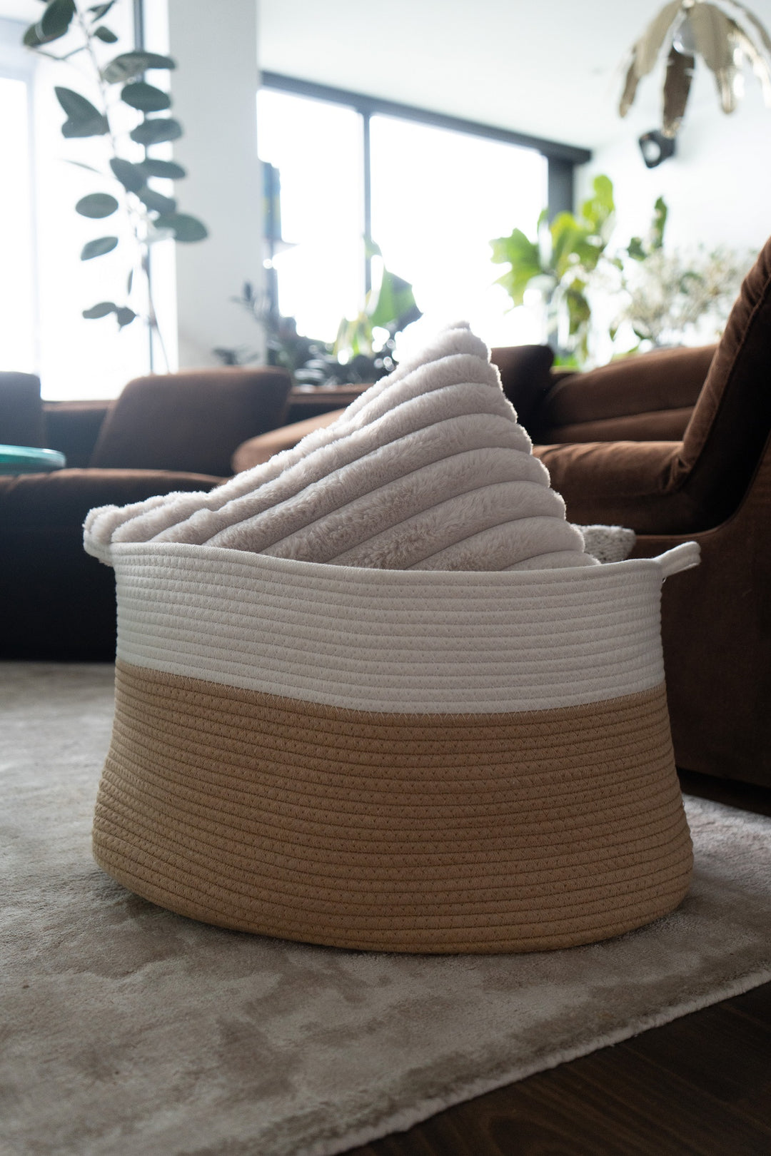 A woven rope storage basket with a two-tone design of beige and white, containing a stack of white towels, set on a rug in a modern living room.